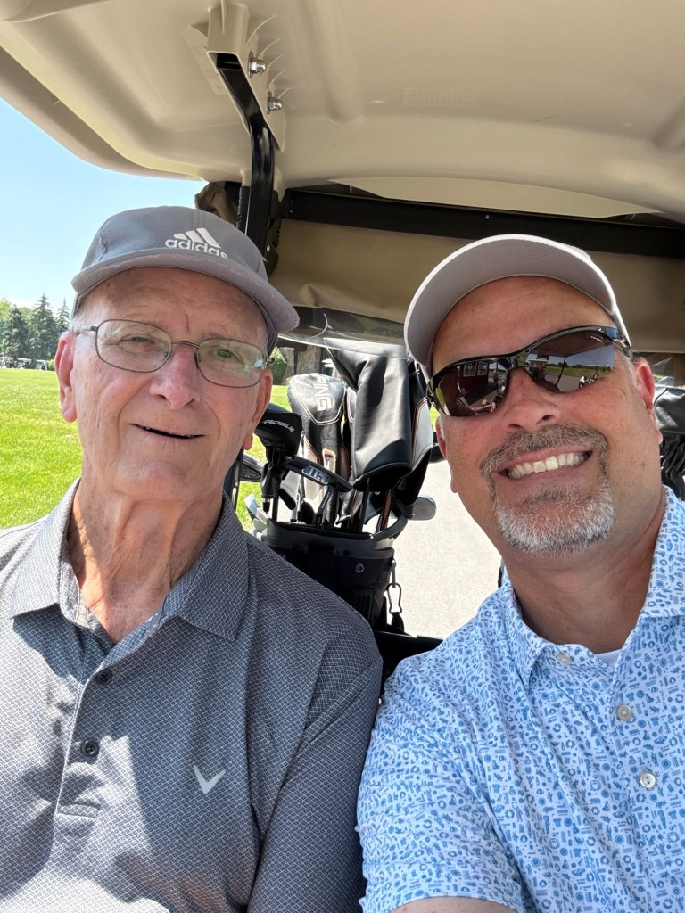 A selfie of two men in a golf cart, smiling. One man is wearing glasses and a gray cap, while the other has a short beard and is wearing sunglasses and a patterned shirt.