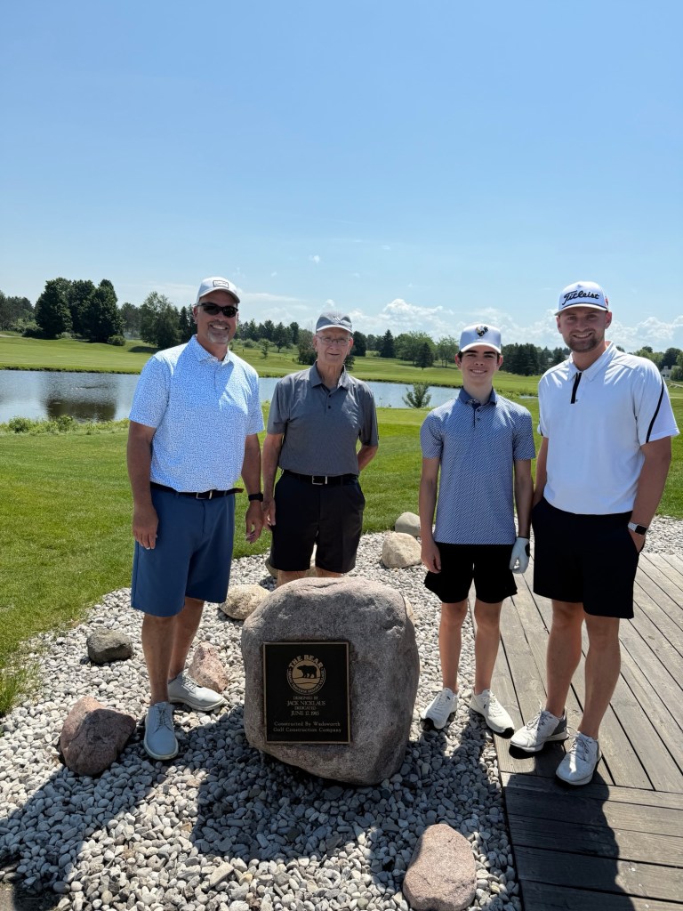 Four men standing together on a golf course near a commemorative stone plaque, with a pond and trees in the background.