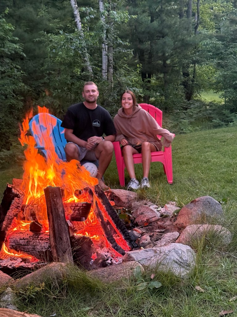 A young couple sitting by a campfire in a forested area, one wearing a black shirt and the other in a brown hoodie, with colorful adirondack chairs in the background.