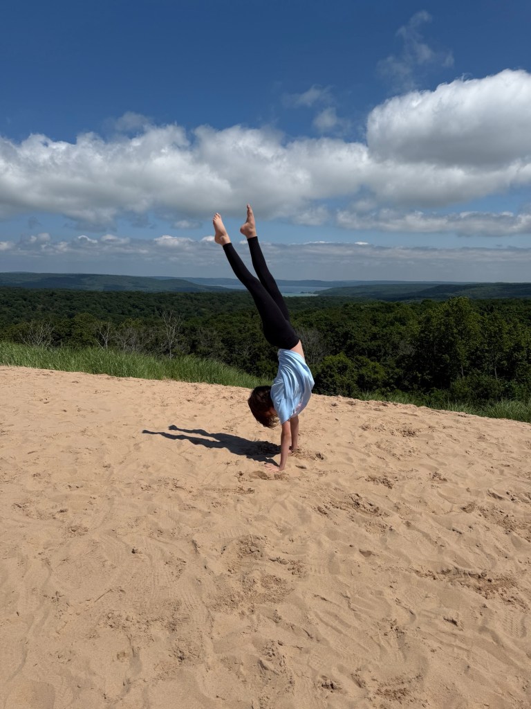 A person performing a handstand on a sandy surface with a scenic view of trees and blue sky in the background.