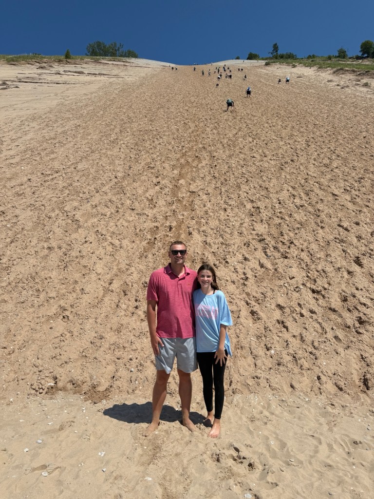 Two individuals stand on a sandy dune with steep slopes in the background, under a clear blue sky.