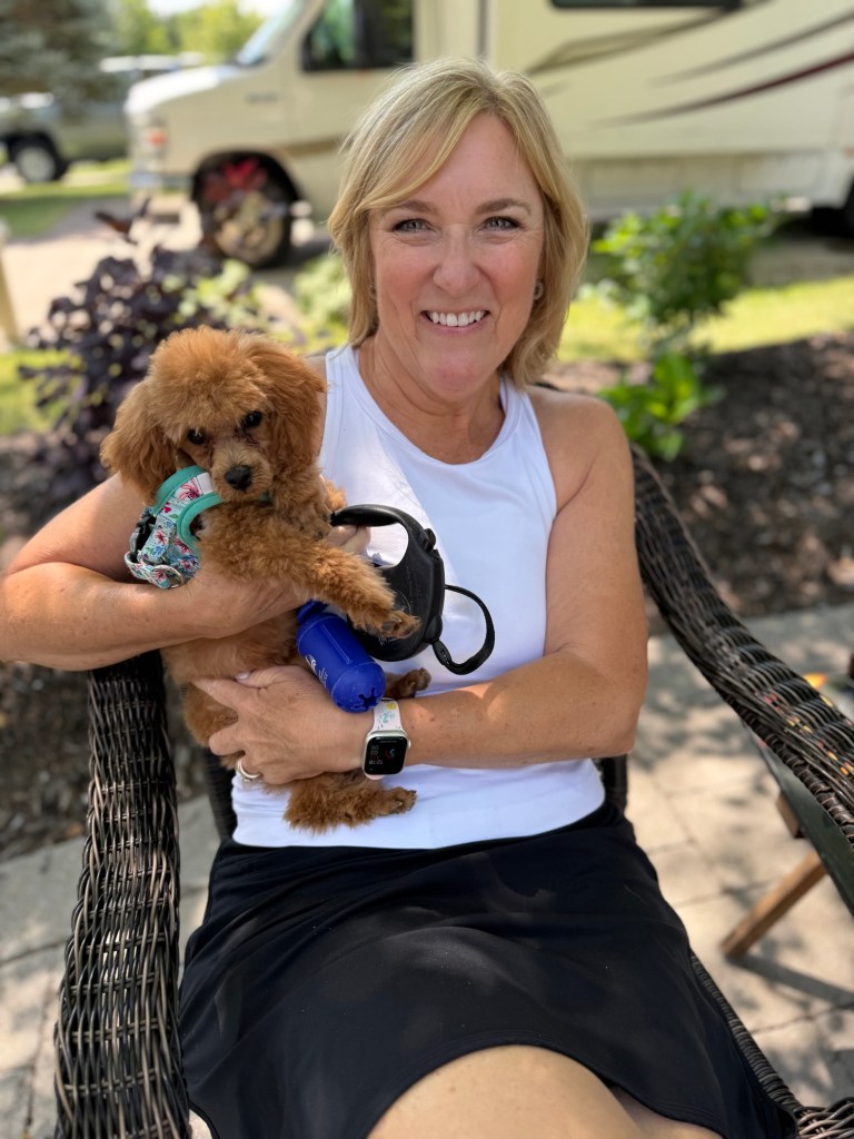 A woman sitting on a chair outside, holding a small toy poodle puppy in her arms, smiling at the camera.