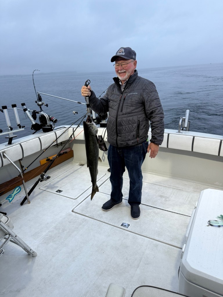 A smiling man holding a freshly caught fish while standing on a fishing boat. The water is calm and the sky is overcast.
