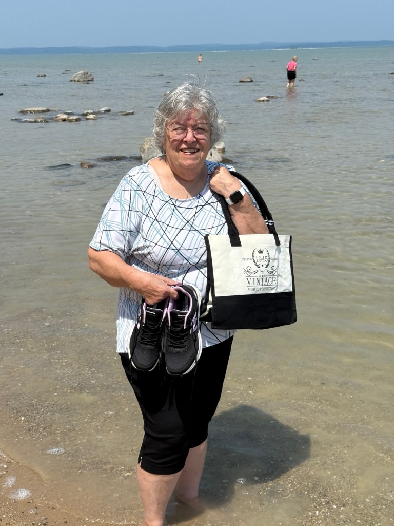 A woman standing in shallow water at the beach, smiling while holding a pair of black shoes and a tote bag.