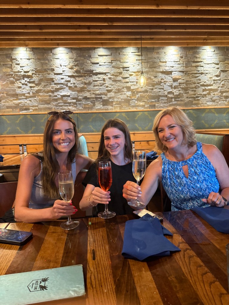 Three women sitting at a table in a restaurant, each holding a drink and smiling at the camera. The interior features wooden accents and stone walls.