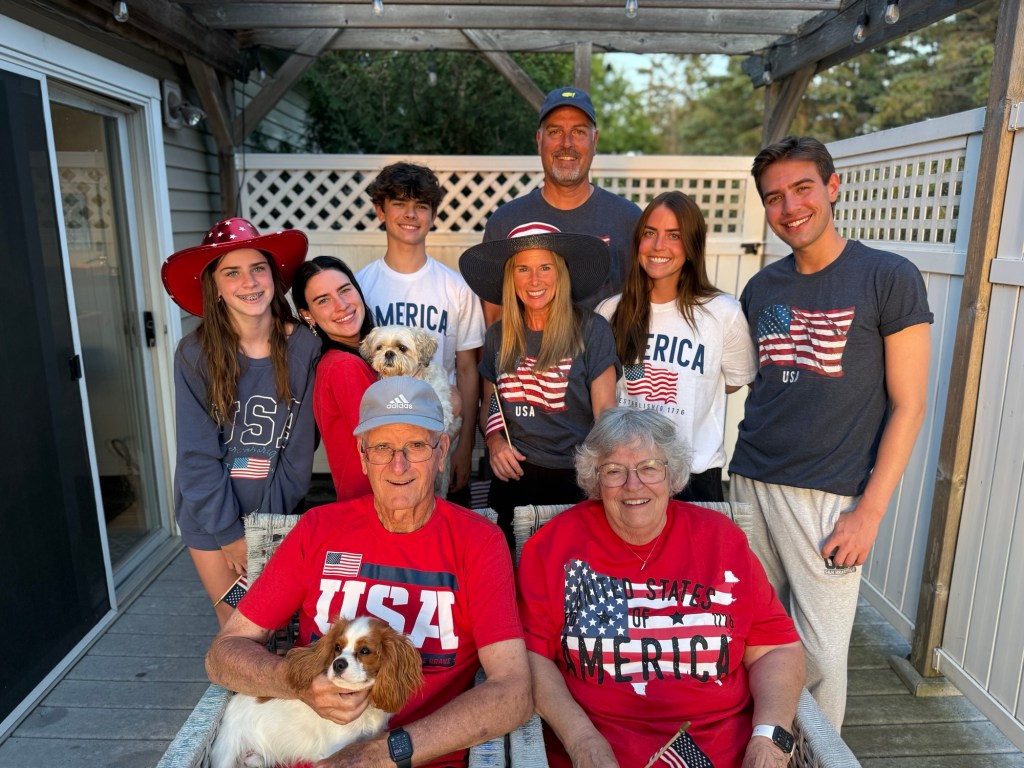 A family photo celebrating togetherness and patriotism, featuring adults and children dressed in red, white, and blue, with some wearing hats and holding a small dog.