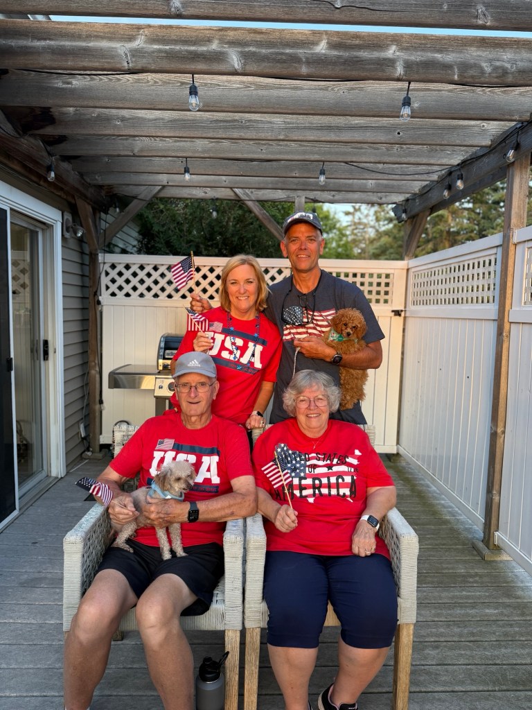 A family photo with four members posing outdoors on a wooden deck, wearing red shirts with patriotic themes and holding small American flags, accompanied by a toy poodle in the man's arms and a small dog on the woman's lap.