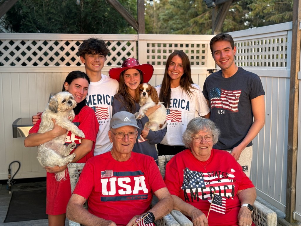A family gathered together outdoors, wearing matching patriotic clothing with American flag designs, smiling and posing with two small dogs.