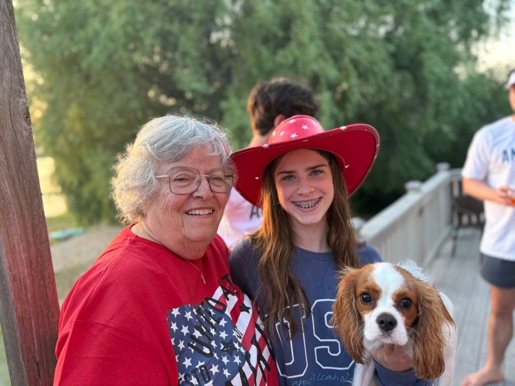 An elderly woman with gray hair and glasses, wearing a red shirt with an American flag, stands next to a teenage girl in a blue shirt with 'USA' printed on it. The girl is wearing a red hat with white stars and is holding a small dog. They are outdoors, smiling, with trees in the background.