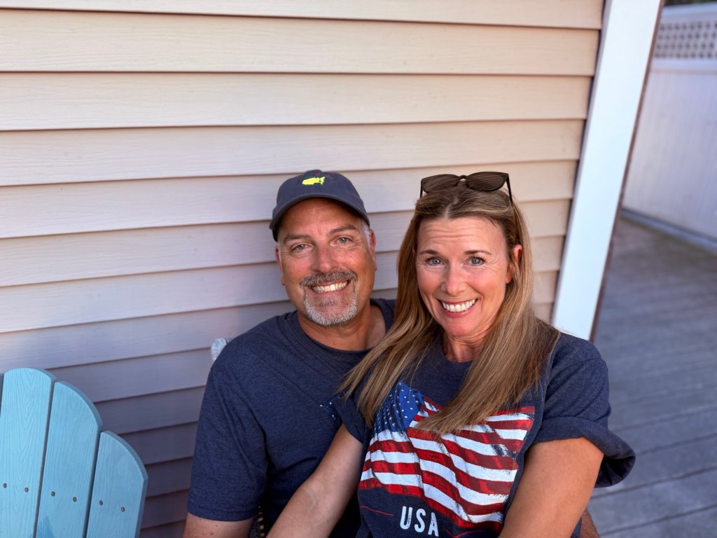 A woman and a man sitting together, smiling, against a wooden wall. The woman wears a shirt with an American flag design, while the man wears a cap and a dark shirt.