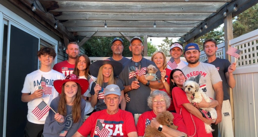 A large family gathering celebrating the 4th of July, with individuals holding small American flags and smiling, surrounded by a rustic wooden structure.