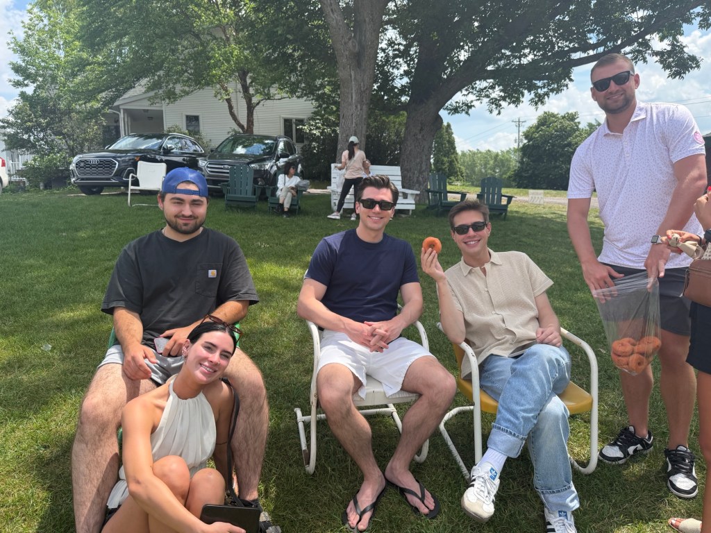 A group of five young adults enjoying a sunny day in a grassy area, sitting on chairs. One person is holding a bag of donuts while others are relaxed and smiling.