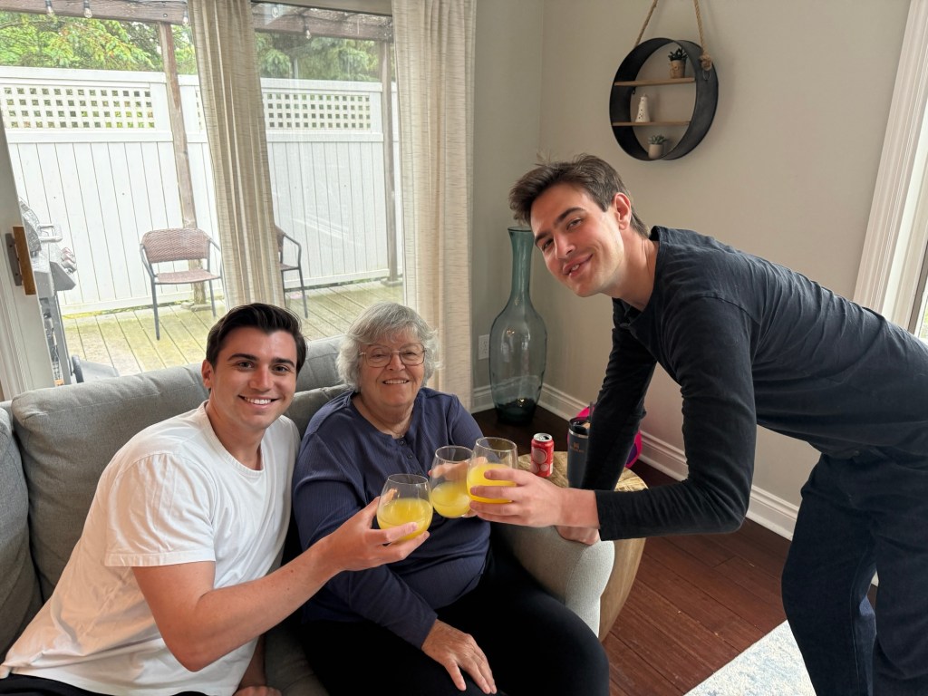 A woman sitting on a couch, holding a drink, surrounded by two young men who are also toasting with their drinks in a cozy living room setting.