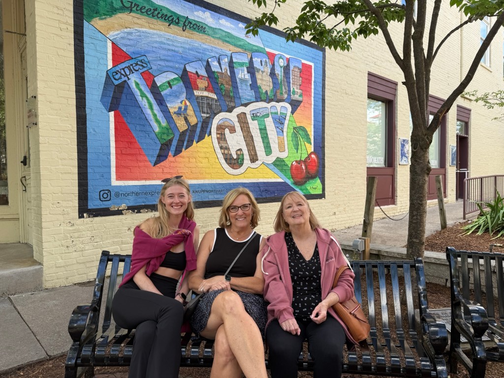 Three women sitting on a bench in front of a colorful mural that says 'Greetings from Traverse City.' The mural features vibrant colors and cherry motifs, representing the local culture.