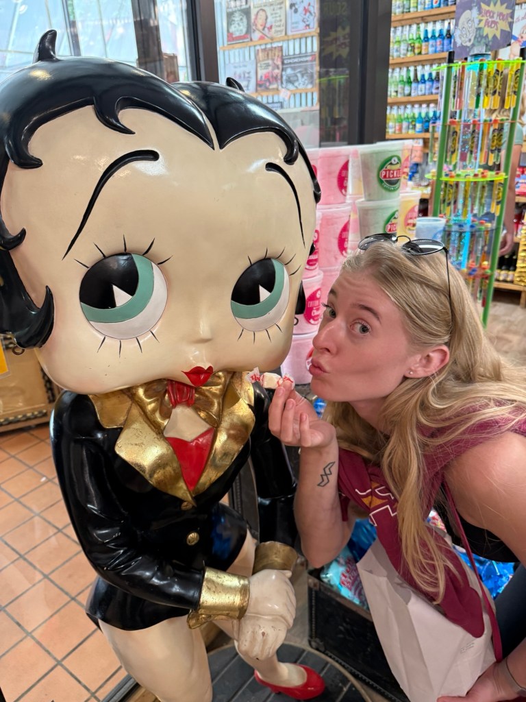 A woman playfully poses with a vintage cartoon character statue while holding a piece of candy, showcasing a fun moment in a colorful store.