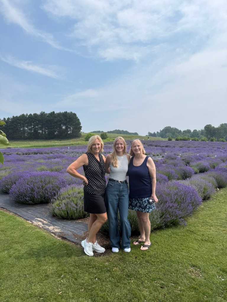 Three women smiling together in a lavender field, with purple lavender plants stretching behind them under a blue sky.