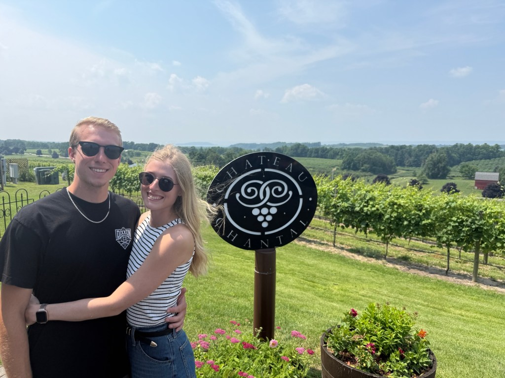 A young couple posing together in front of a vineyard sign, with a scenic landscape of vineyards and rolling hills in the background.