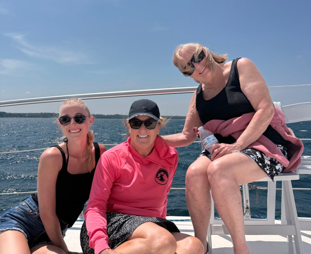 Three women smiling and enjoying a sunny day on a boat, with a lake in the background.