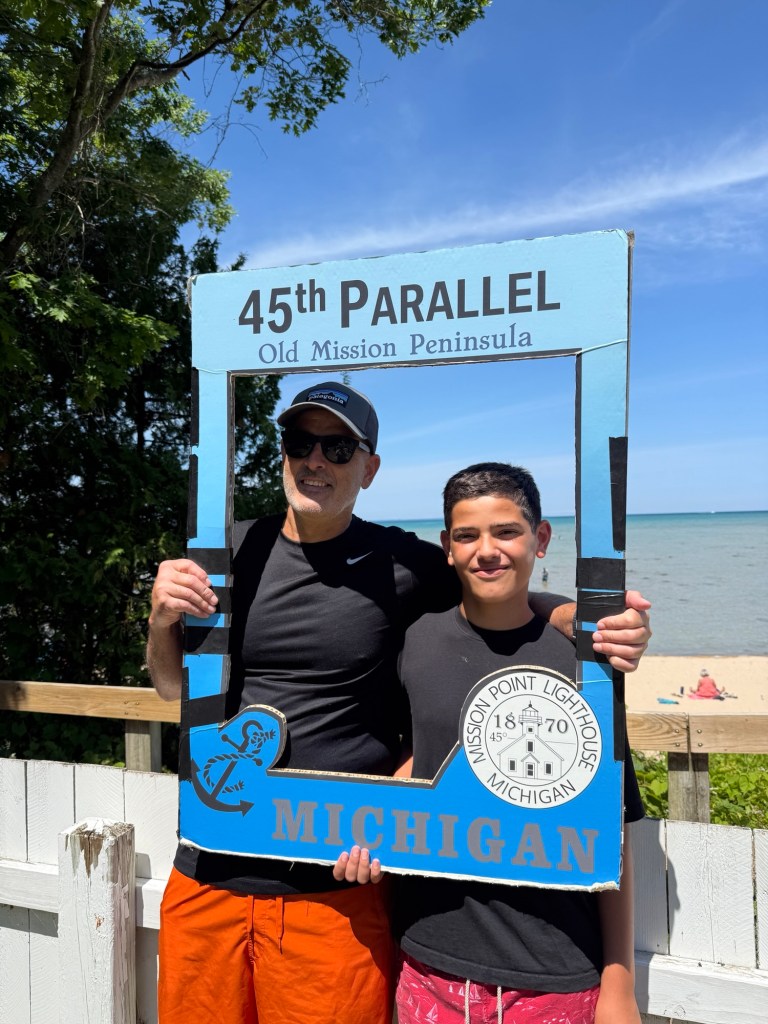 Two men posing together inside a decorative frame that reads '45th Parallel Old Mission Peninsula Michigan' with a beach and water in the background.