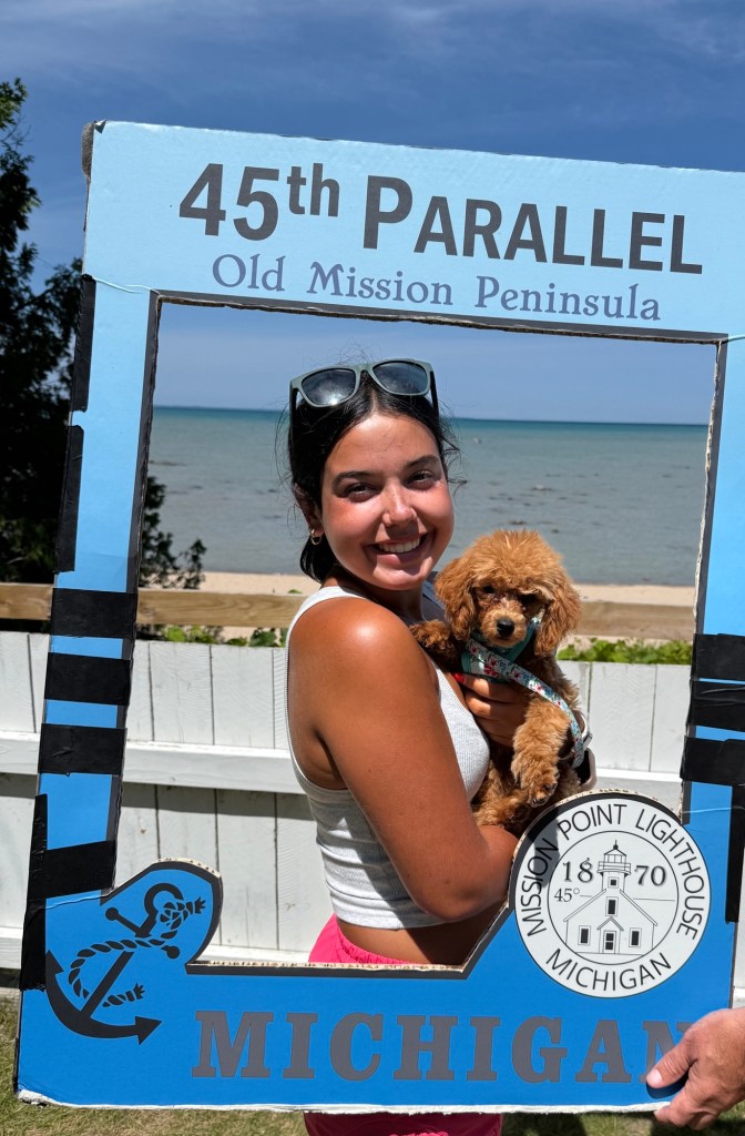 A young woman holding a small brown puppy, posing in front of a sign reading '45th Parallel, Old Mission Peninsula' with a scenic view of the lake in the background.