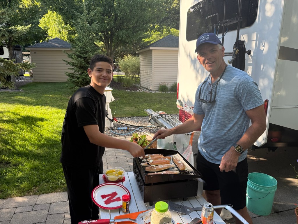 A man and a boy grilling fish outdoors next to a camper, with a lush green background.