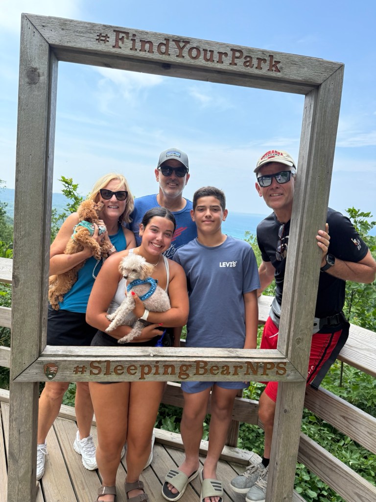 A family poses together in a wooden photo frame at Sleeping Bear Dunes National Lakeshore, with two small dogs in their arms and a scenic view in the background.