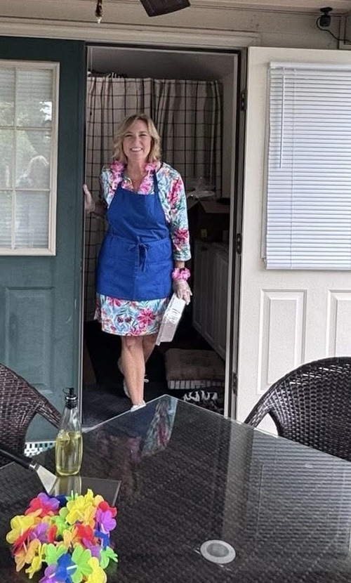 A smiling woman wearing a blue apron and a colorful floral dress stands at the doorway of a home, holding a container in her right hand, with a table set outside adorned with a colorful flower lei and a bottle.