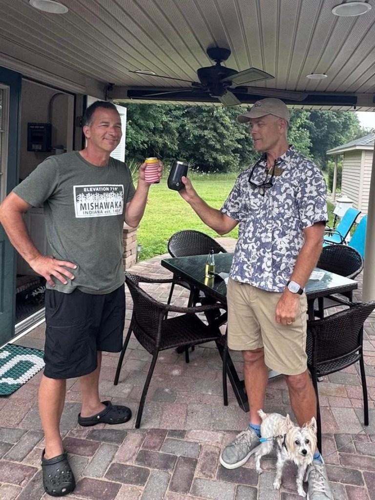 Two men holding drinks while standing on a patio, with a small dog at their feet.
