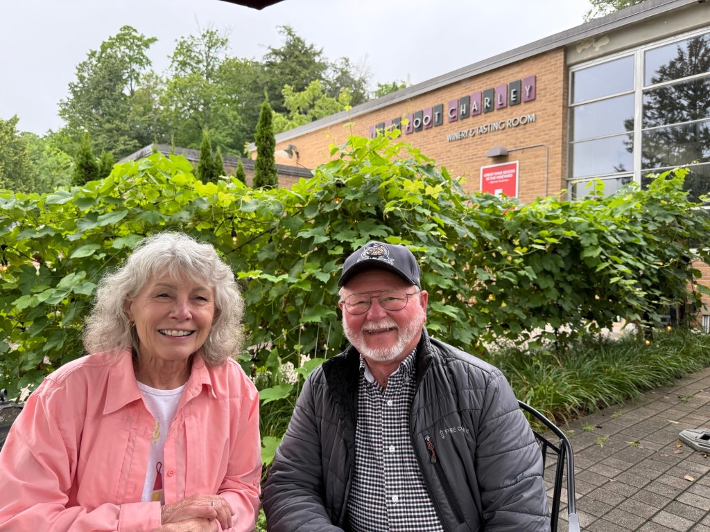 A smiling couple seated at an outdoor table in front of a winery, surrounded by green vines and greenery.