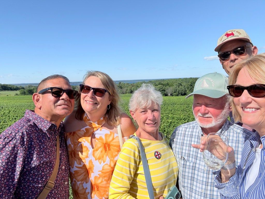 A group of six friends posing for a selfie in a vineyard, smiling and enjoying a sunny day with fields of grapevines in the background.