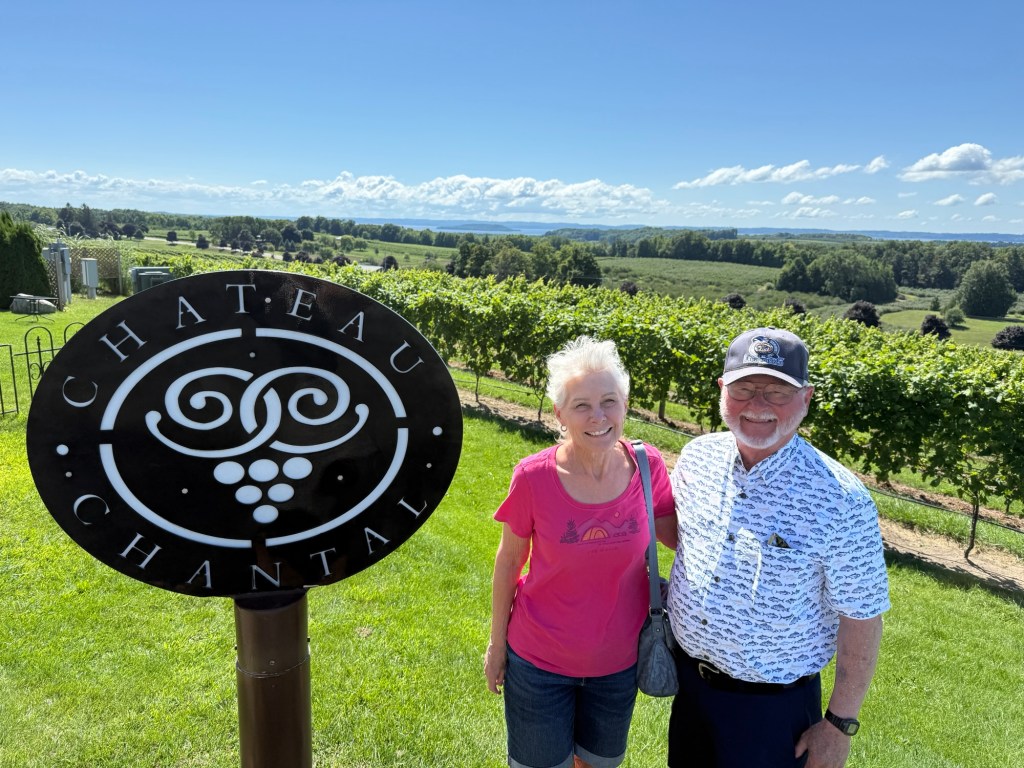A couple stands smiling in front of the Chateau Chantal vineyard sign, with lush green vineyards and a clear blue sky in the background.