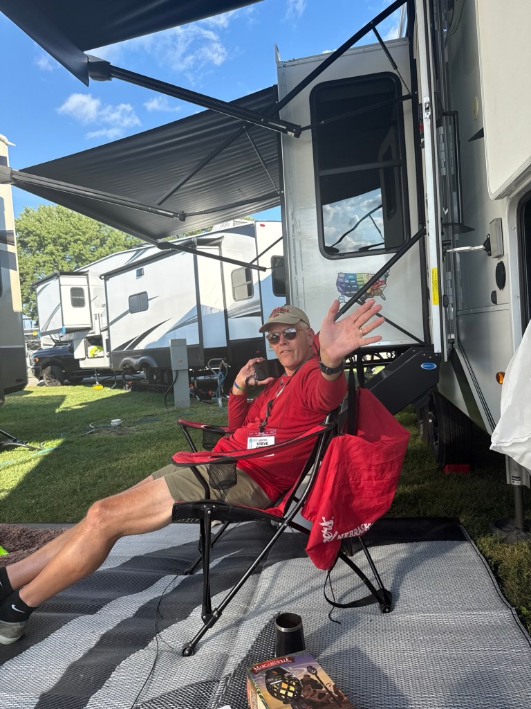 A man sitting in a camping chair outside a trailer, waving and smiling, with a book and a cup on the ground beside him.