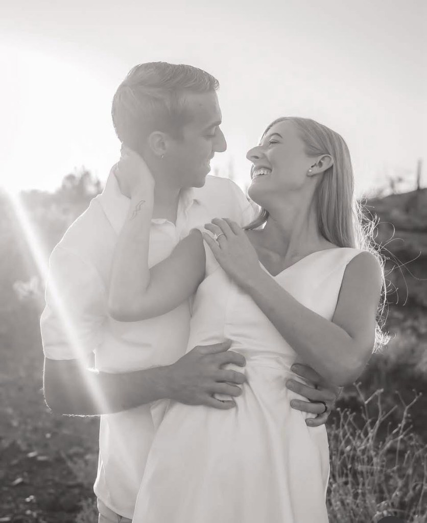 A couple smiling and embracing each other in a sunlit outdoor setting, captured in black and white.