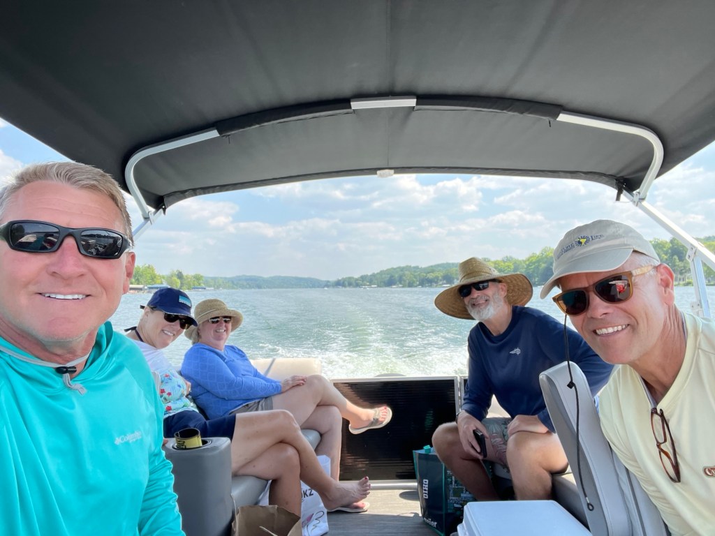 A group of five people enjoying a day on a boat in a scenic lake, with trees and hills visible in the background.