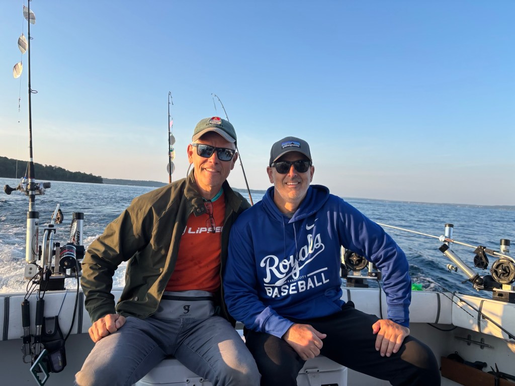 Two men sitting on a fishing boat with fishing rods in the background, enjoying a sunny day on the water.