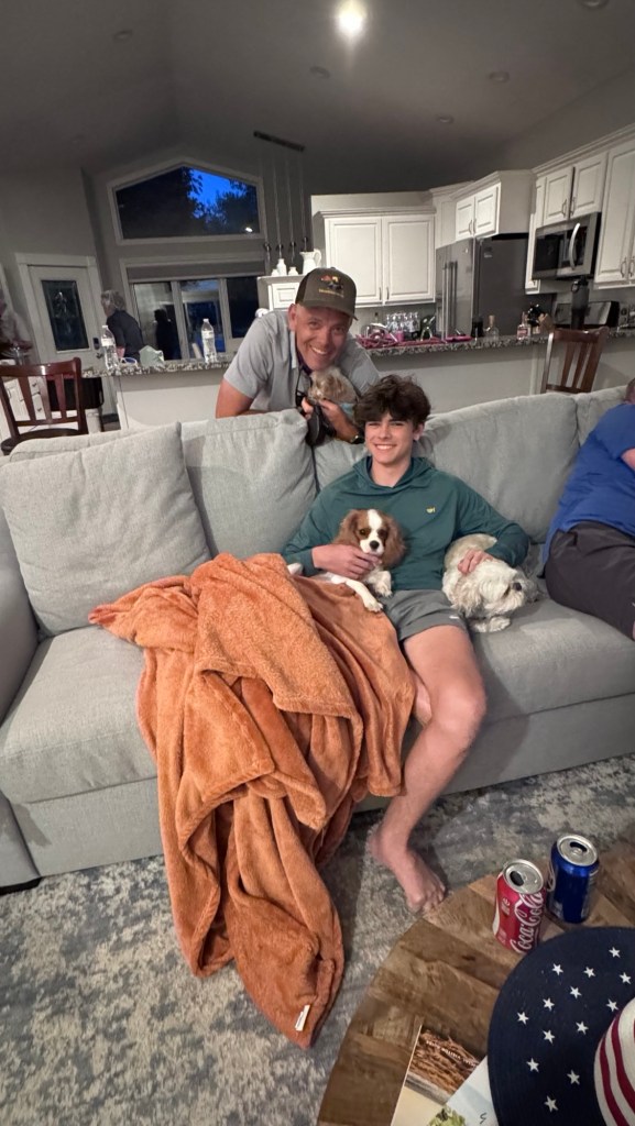 A man and a teenager sitting on a couch with two small dogs, one of which is being held by the man. The couch is covered with an orange blanket, and there are soft drinks on a nearby table.