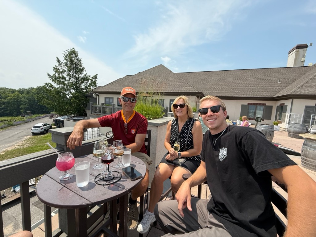 A group of three people sitting at a wooden table on a sunny day, enjoying drinks at a winery. The background features trees and a building.