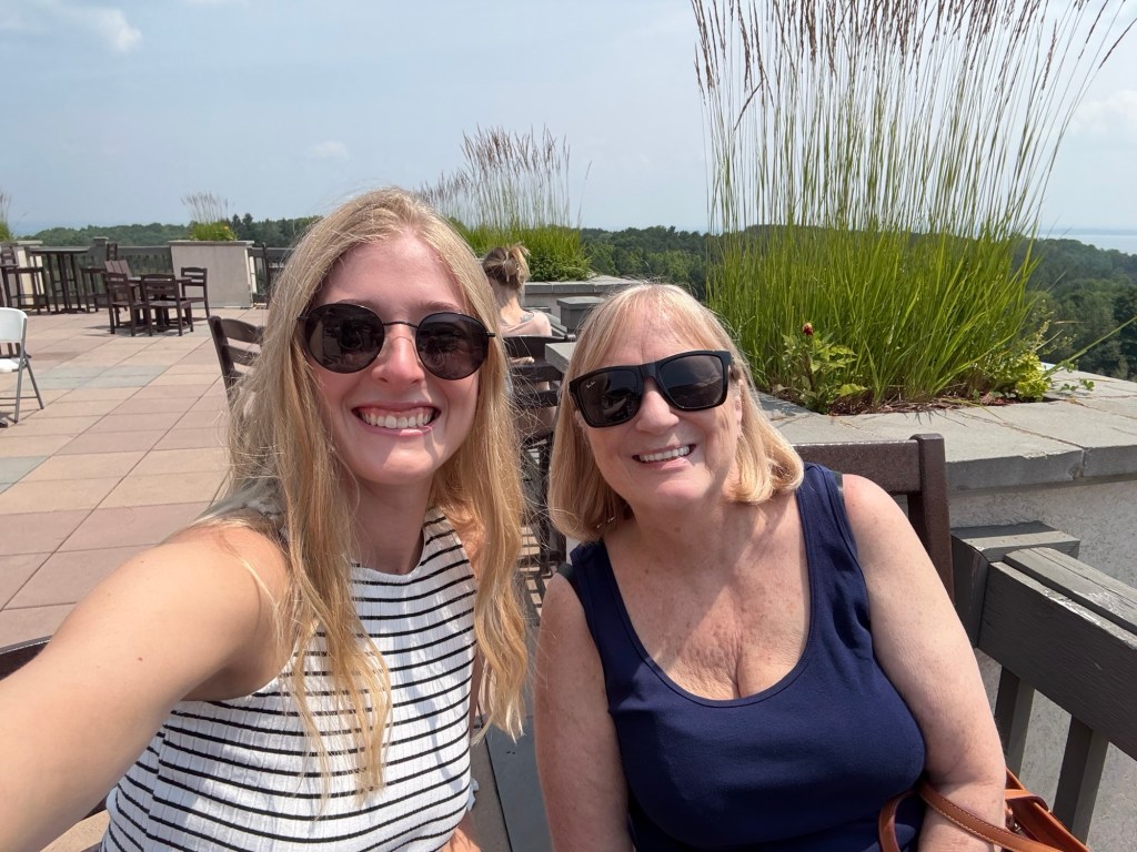 Two women smiling for a selfie outdoors, wearing sunglasses. They are seated on a patio with grass and trees in the background.