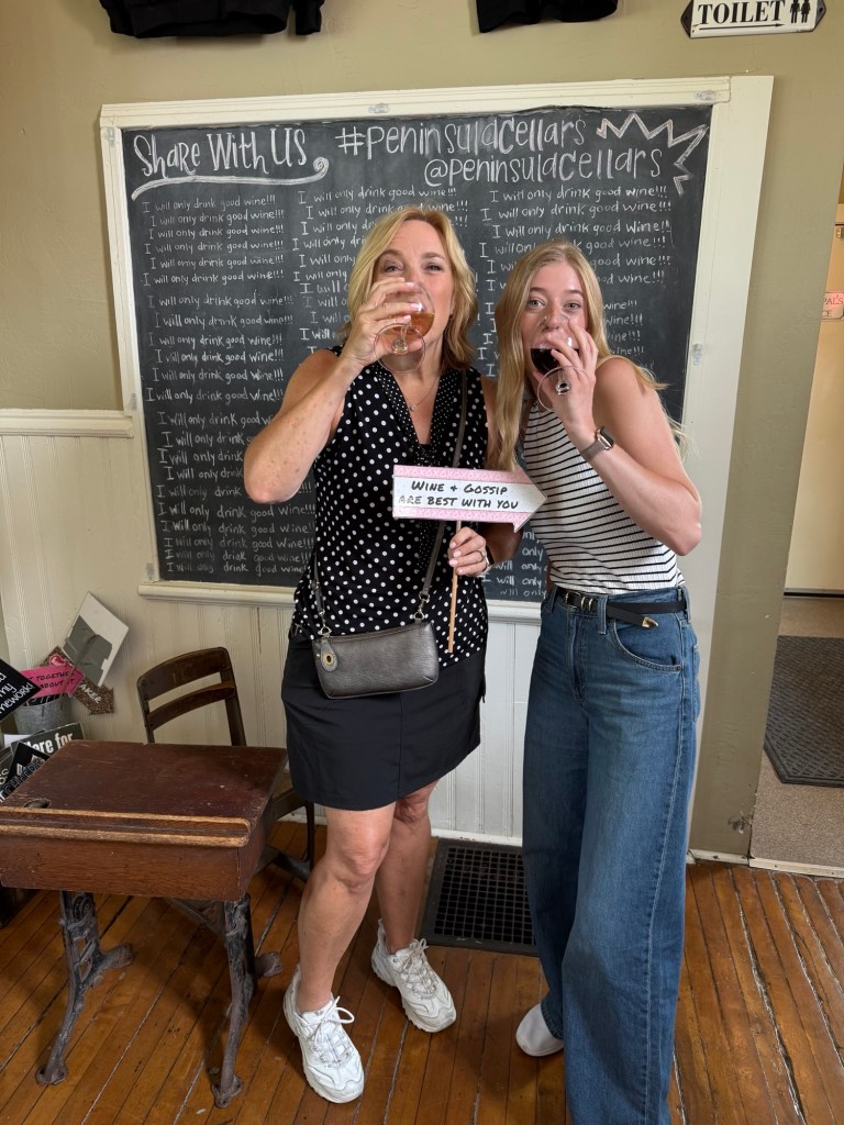 Two women enjoy wine tasting at a vineyard, holding a sign that reads 'Wine + Gossip are best with you'. The background features a chalkboard with wine-related text.