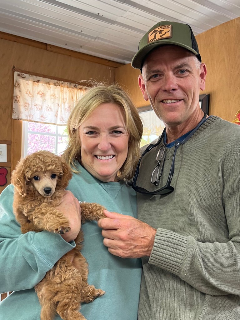 A smiling couple holds a small toy poodle puppy in a cozy indoor setting.