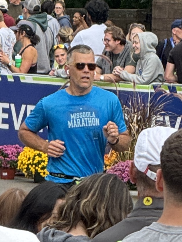 A male runner in a blue t-shirt with 'Missoula Marathon' printed on it, wearing sunglasses and earbuds, running past a crowd of spectators enjoying flowers in pots.