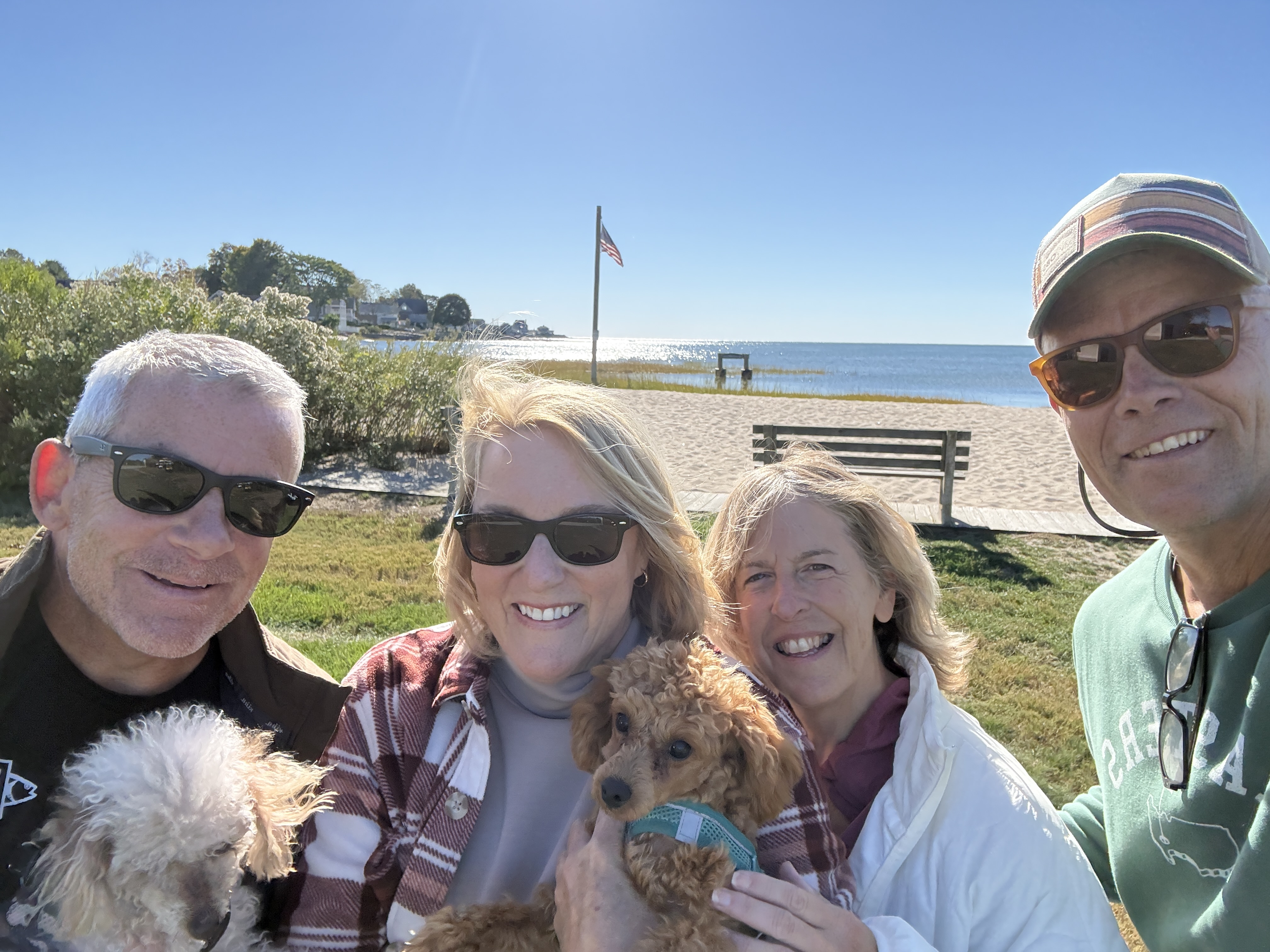 A group of four adults smiling outdoors, holding two small dogs. They are wearing sunglasses and standing on a grassy area with a beach in the background. An American flag is visible, and the sky is clear and sunny.