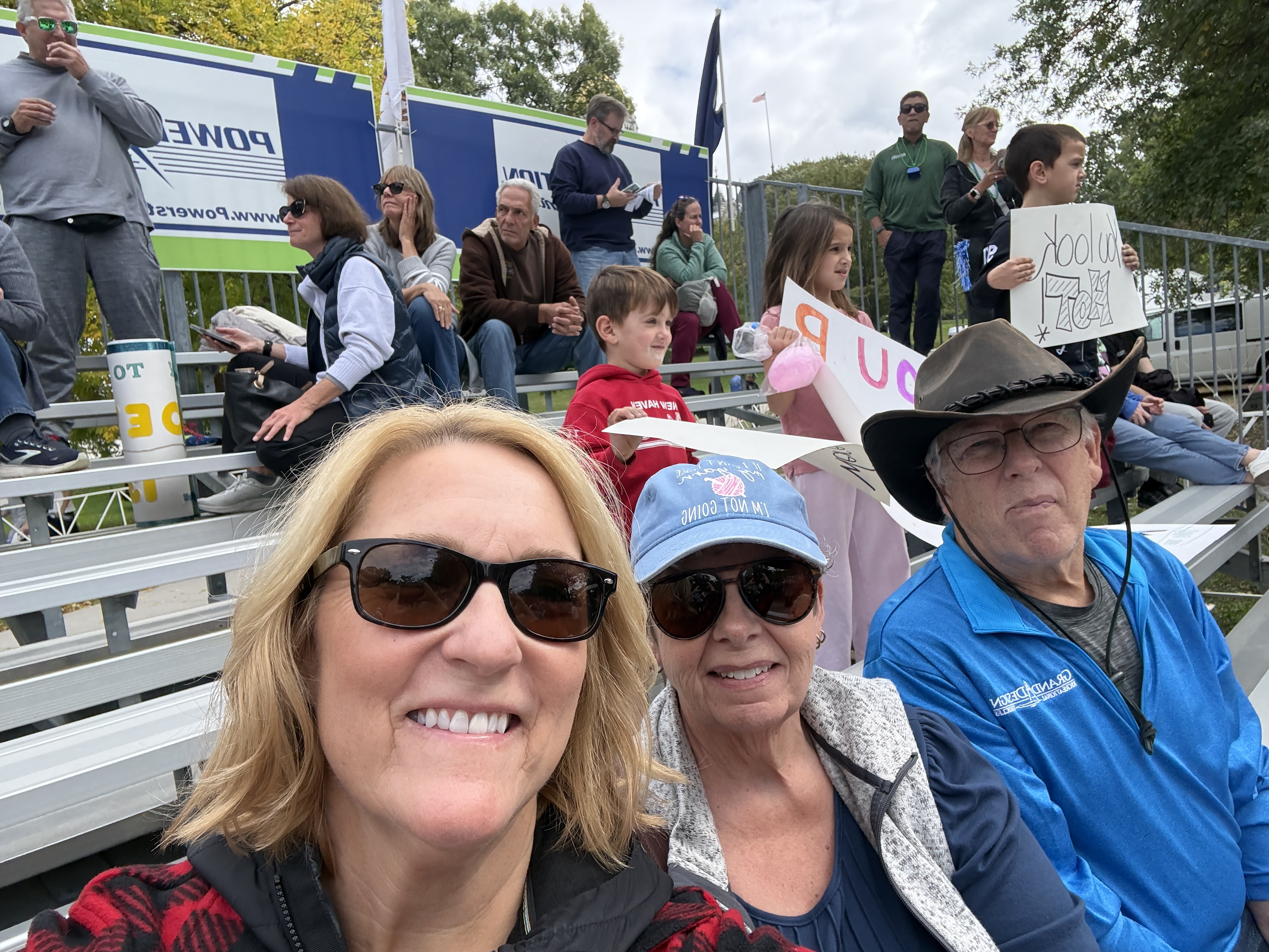 A group of people sitting in bleachers, with three adults in the foreground taking a selfie. One woman is wearing sunglasses and a baseball cap, while another is wearing a gray jacket. An older man with glasses is sitting next to her. The background shows several spectators, including children holding signs.