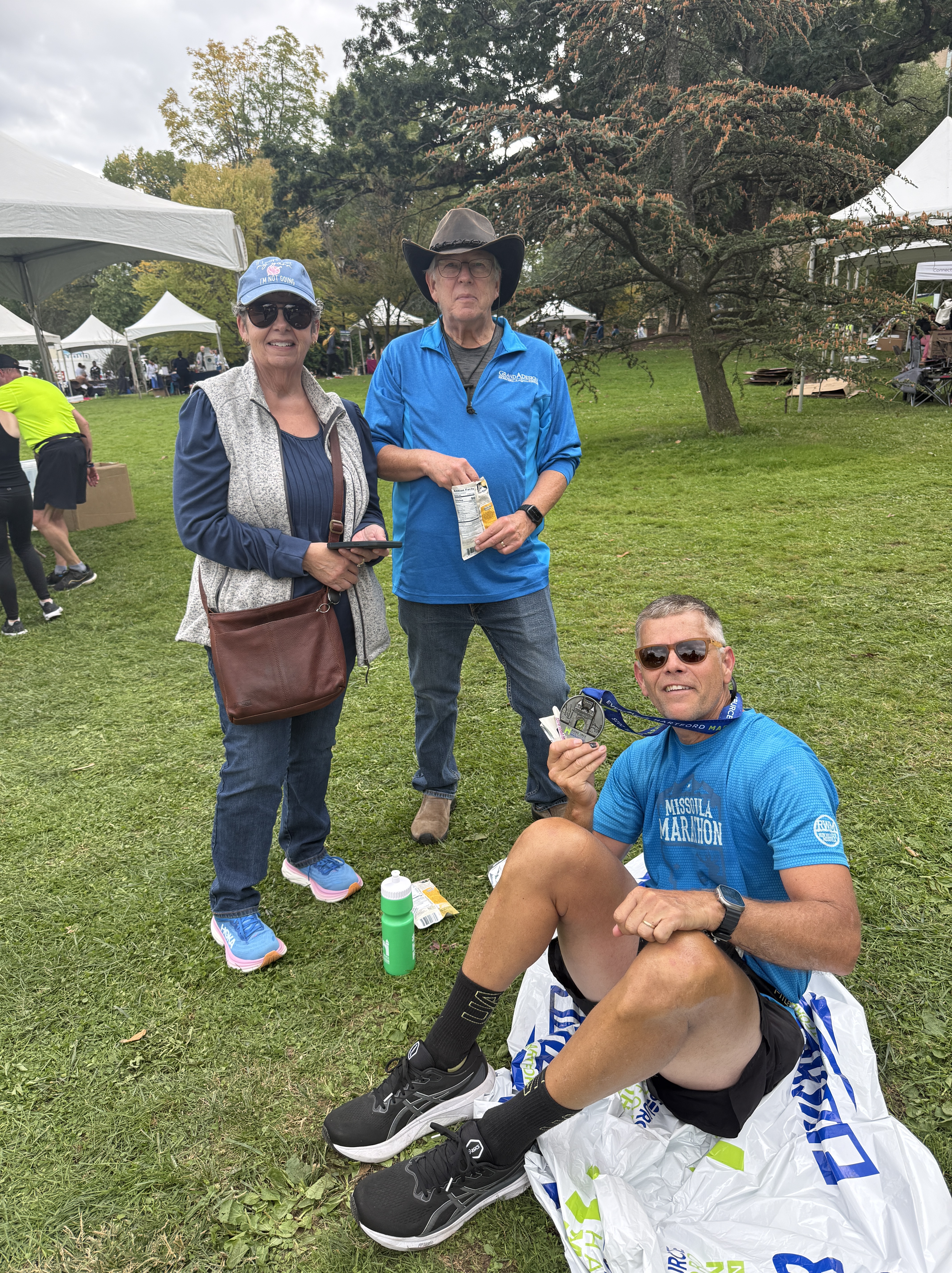 Three individuals at an outdoor event: one woman standing with sunglasses and a hat, a man in a blue shirt holding a snack, and another man sitting on the grass displaying a marathon medal.