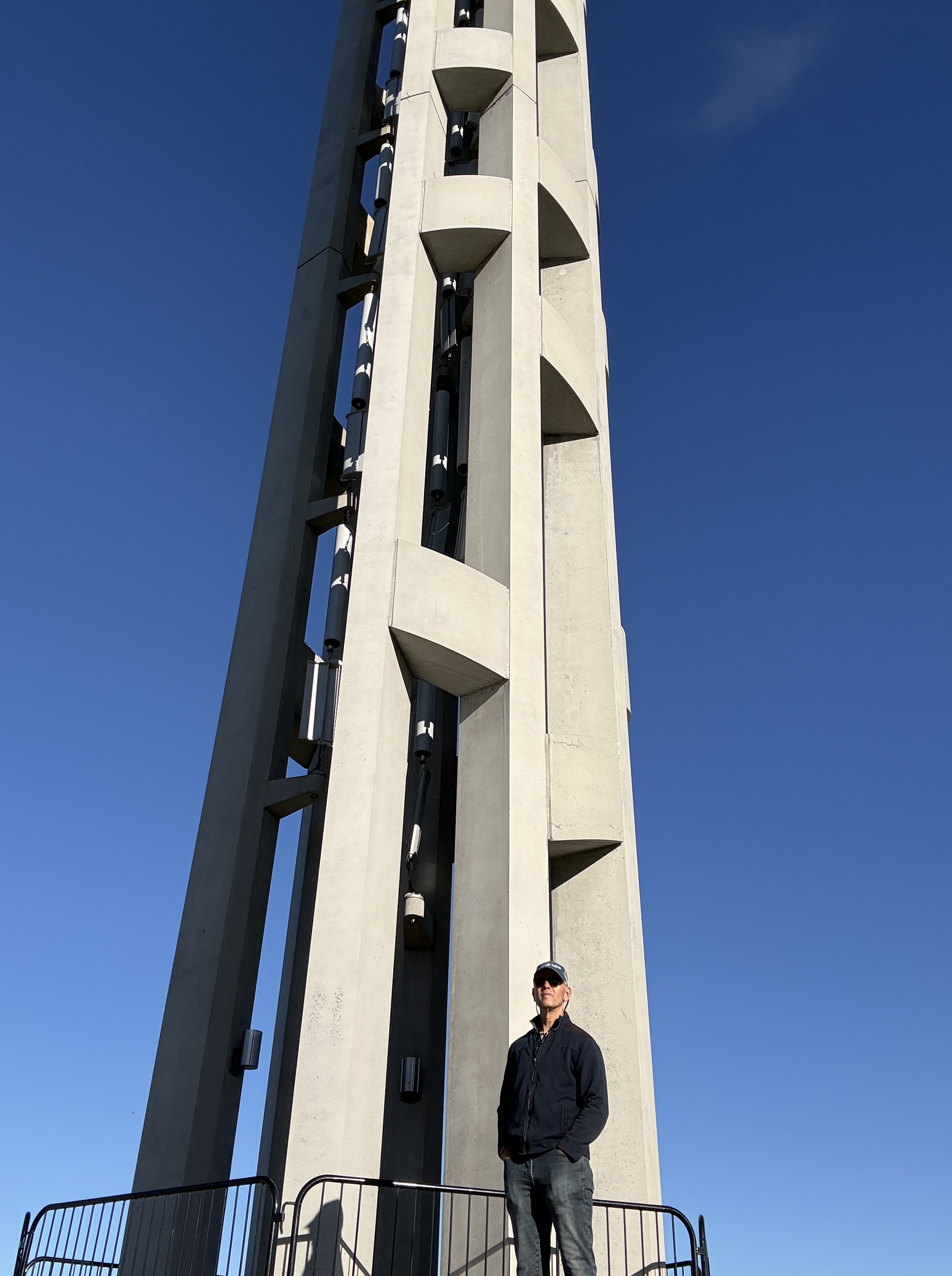 A person standing near a tall, modern concrete tower against a clear blue sky.