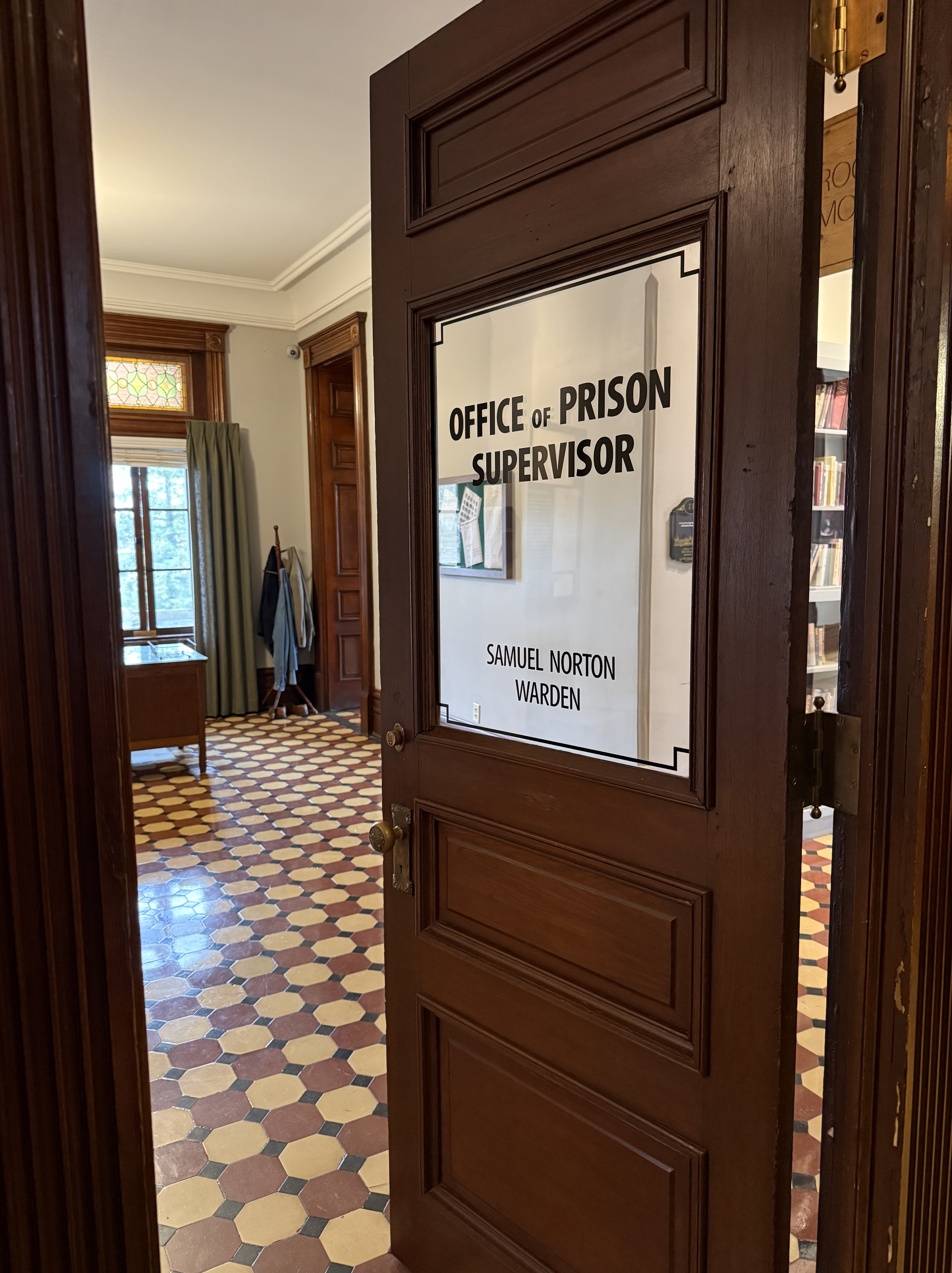 A wooden door with a glass panel displaying the text 'OFFICE of PRISON SUPERVISOR' and 'SAMUEL NORTON WARDEN'. The view shows a room behind the door with patterned tiled flooring, a wooden table, and curtains.
