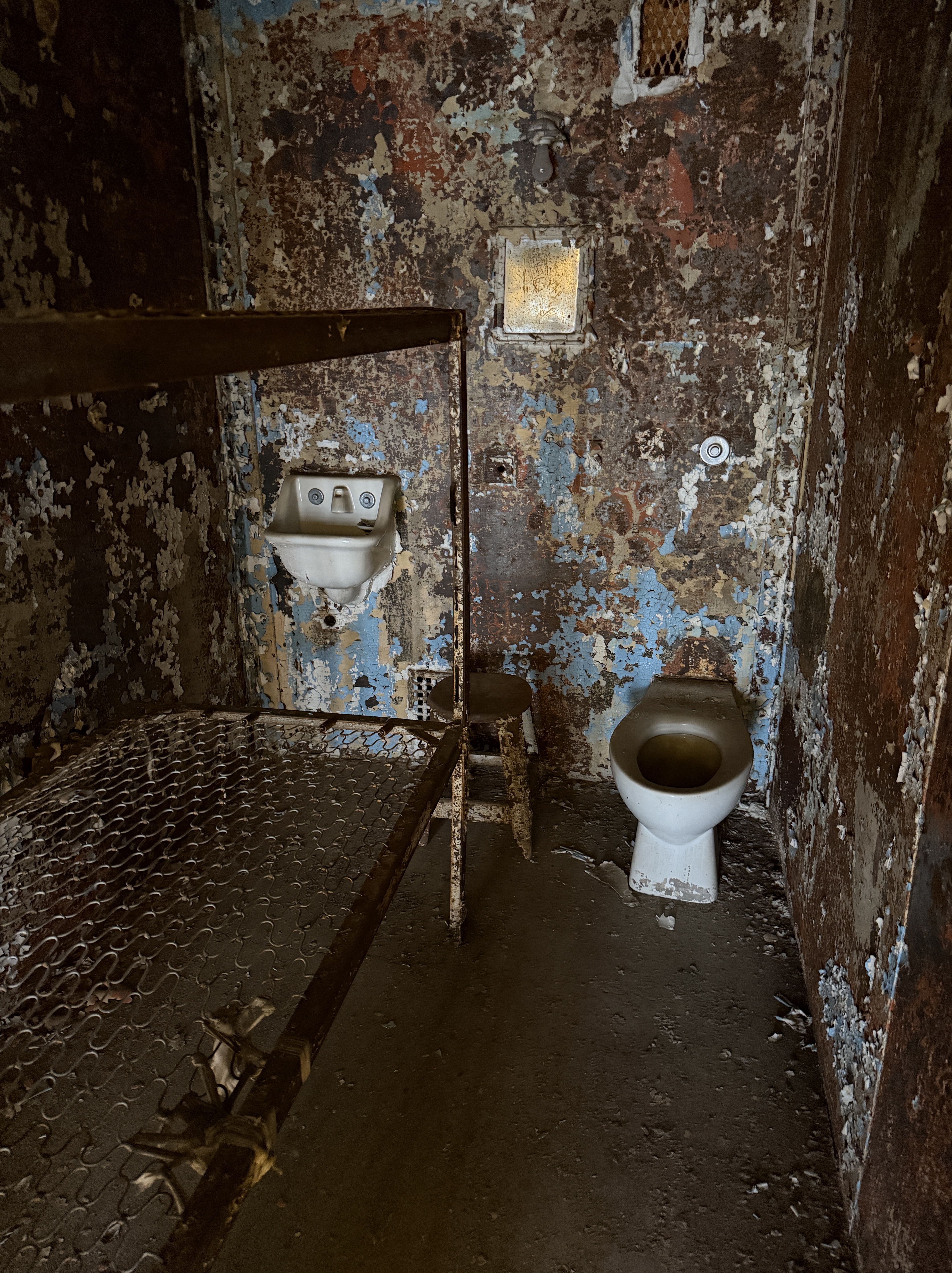 Interior of an abandoned prison cell featuring a rusty metal bed frame, a sink, and a toilet on a concrete floor with peeling paint on the walls.