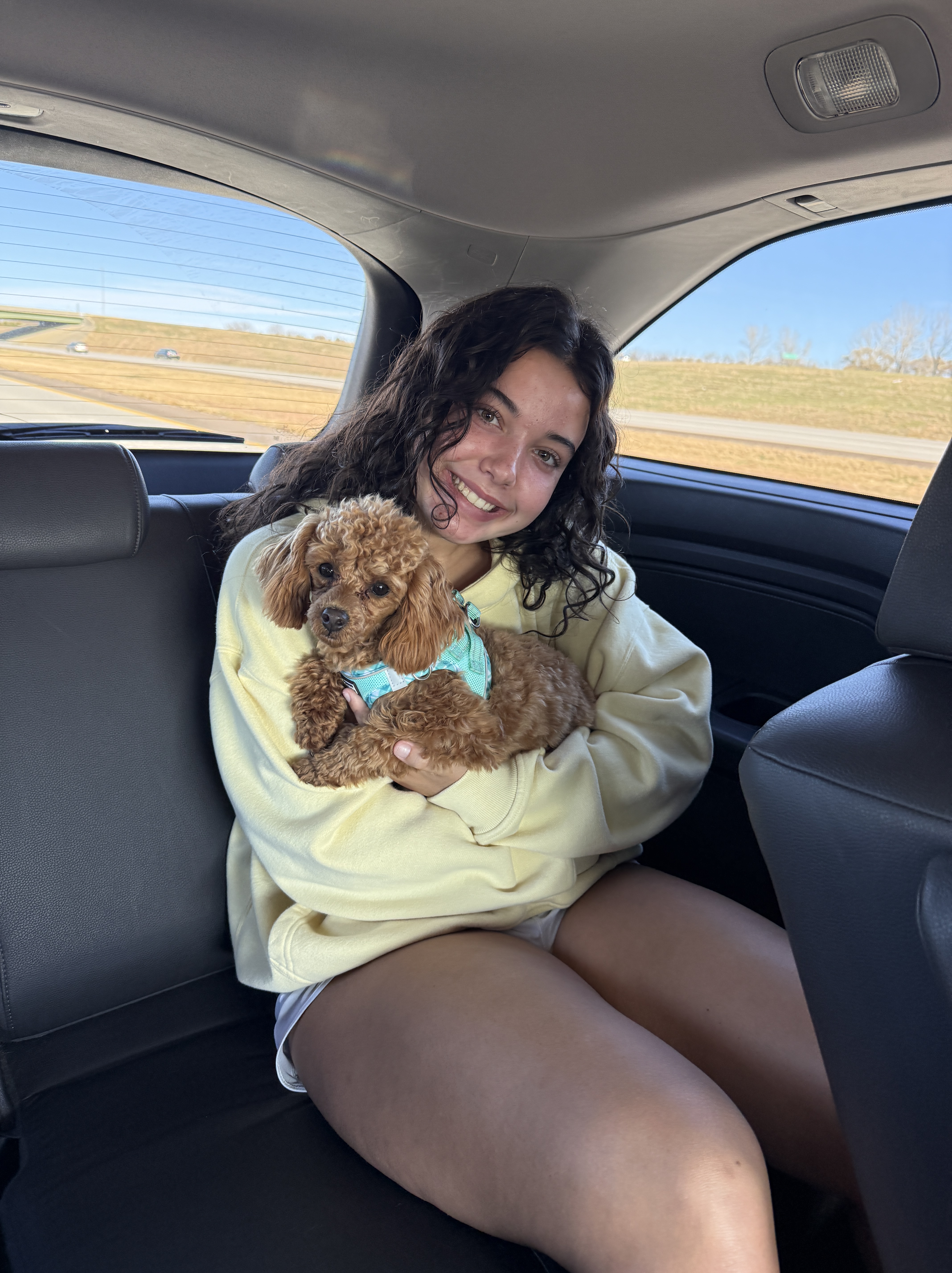 A young woman sitting in the backseat of a car, smiling while holding a small brown dog with curly fur, wearing a turquoise harness. The background shows a clear blue sky and grassy fields outside the car window.