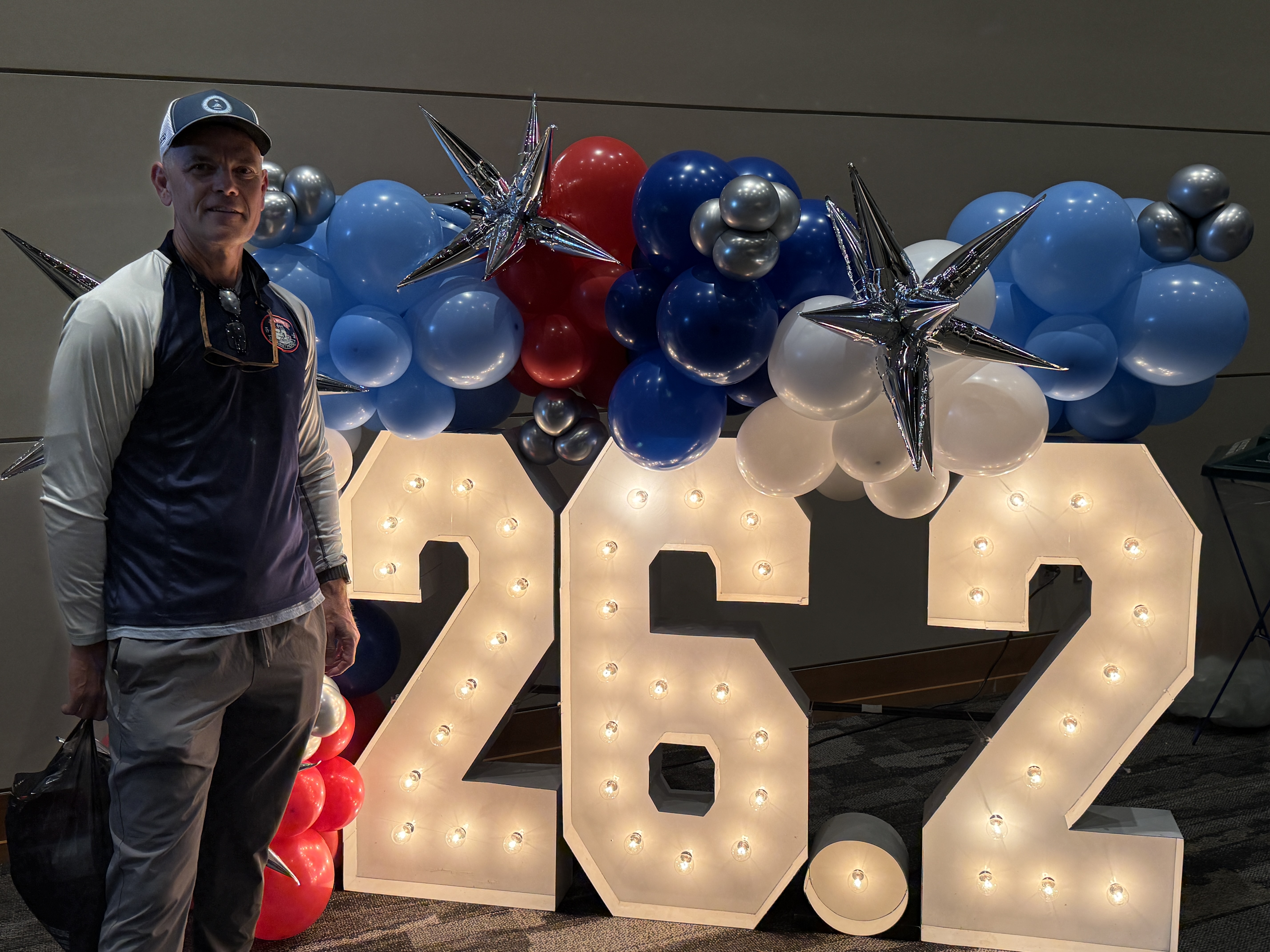 A man poses next to a large illuminated sign that reads '26.2', surrounded by colorful balloons and star decorations.