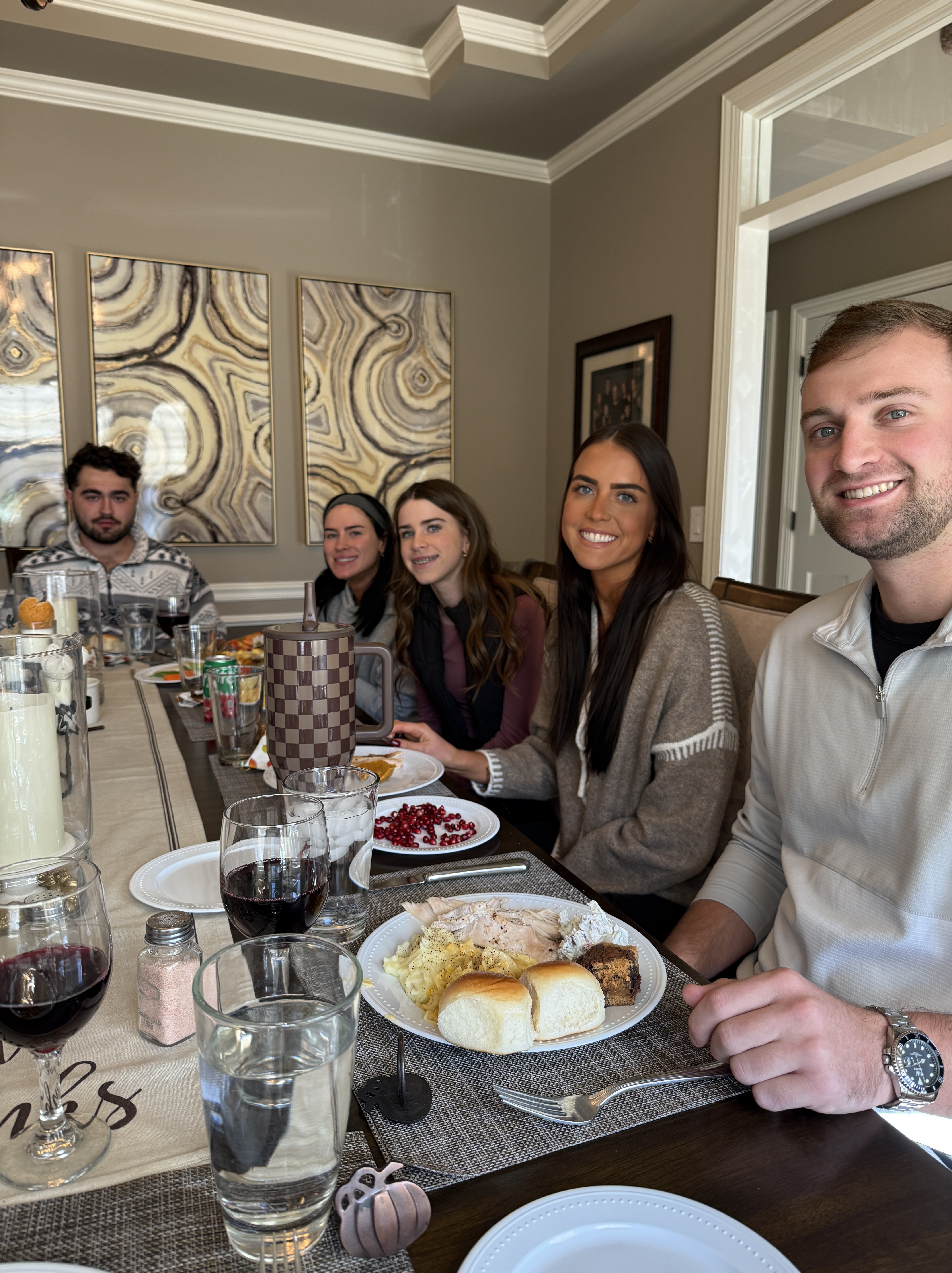 A group of five people sitting at a dinner table with plates of food, drinks, and a festive atmosphere. The table is set with various dishes, including turkey and rolls, and decorative elements are visible in the background.